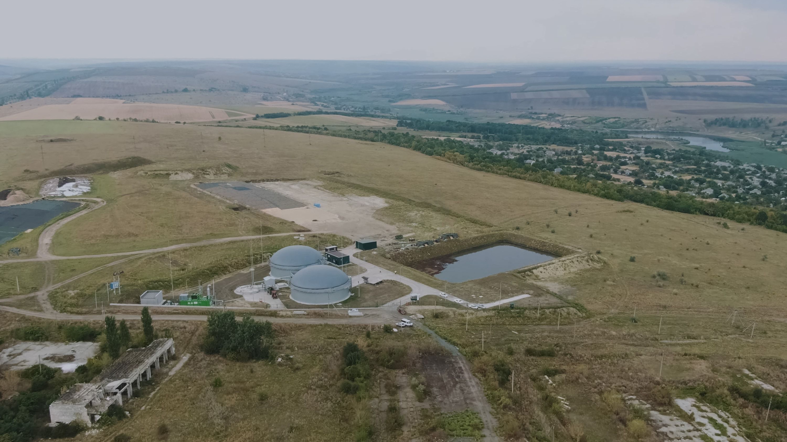 Vista aérea de una planta de biogás con dos digestores y una balsa, en medio de un amplio paisaje agrícola con un pueblo y un río al fondo