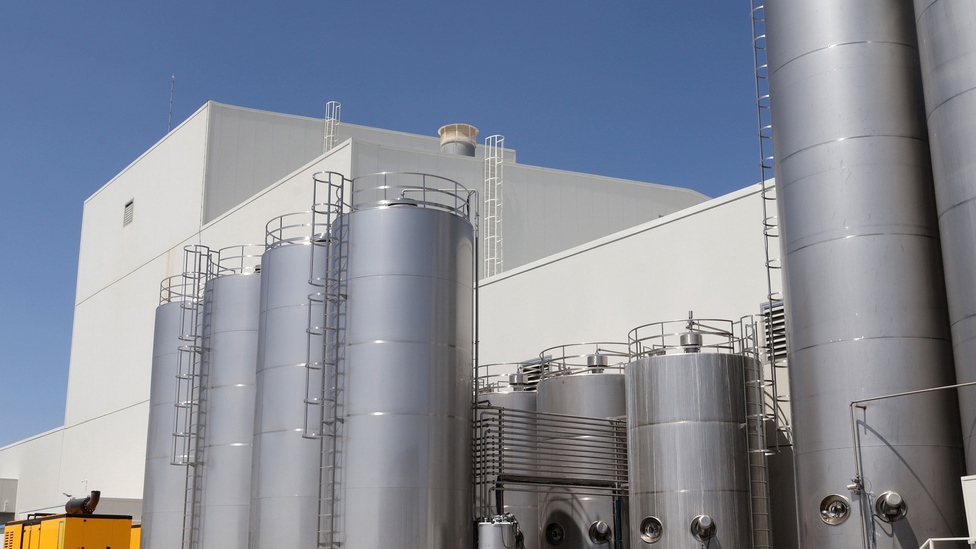 Exterior de una planta agroalimentaria con depósitos verticales de acero inoxidable y sus escaleras de acceso junto a la nave de producción, bajo un cielo despejado