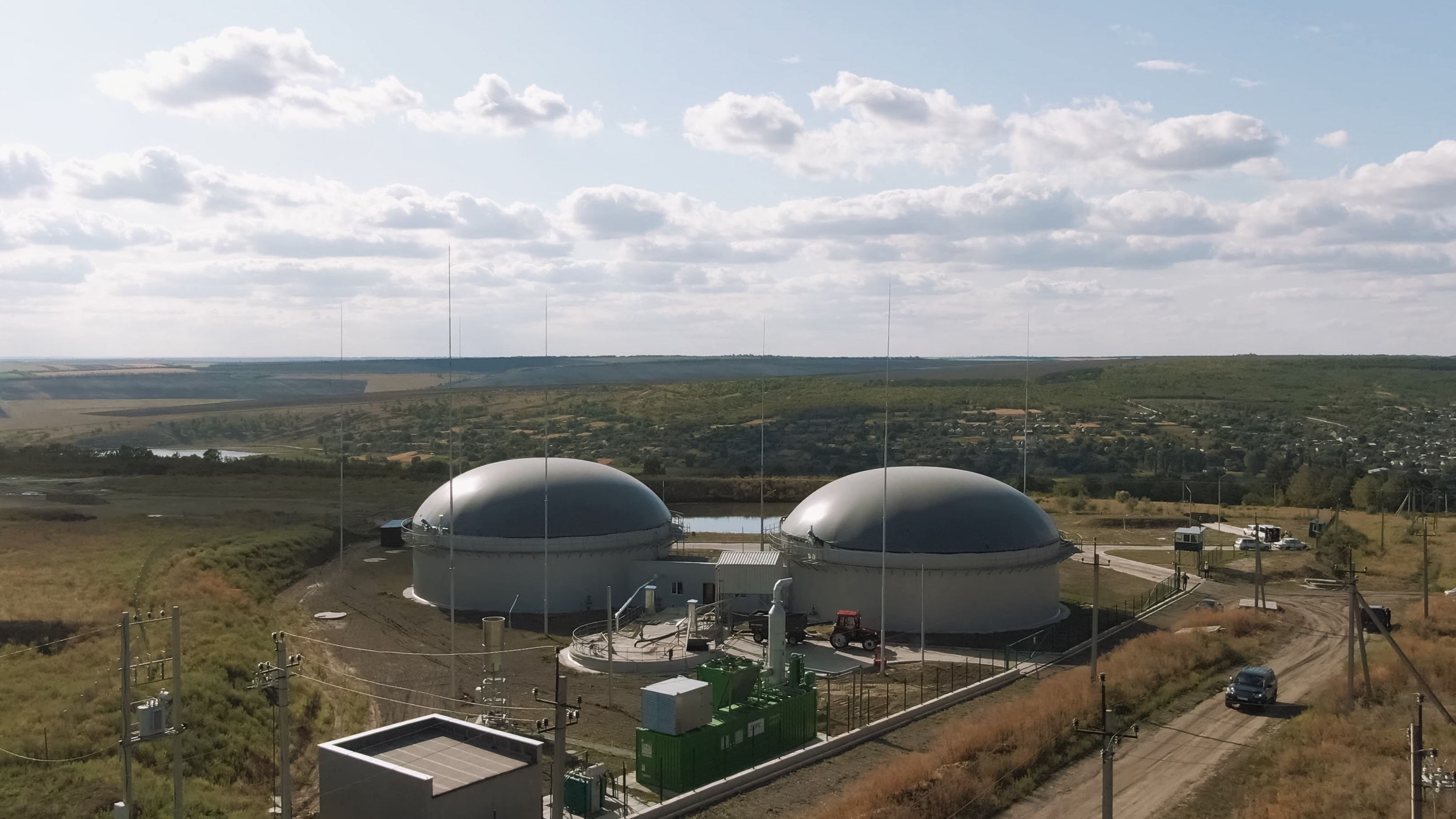 Planta de biogás en entorno agrícola con dos digestores de cubierta de membrana gris, el contenedor verde del motor de cogeneración con su chimenea y una caseta técnica en primer plano
