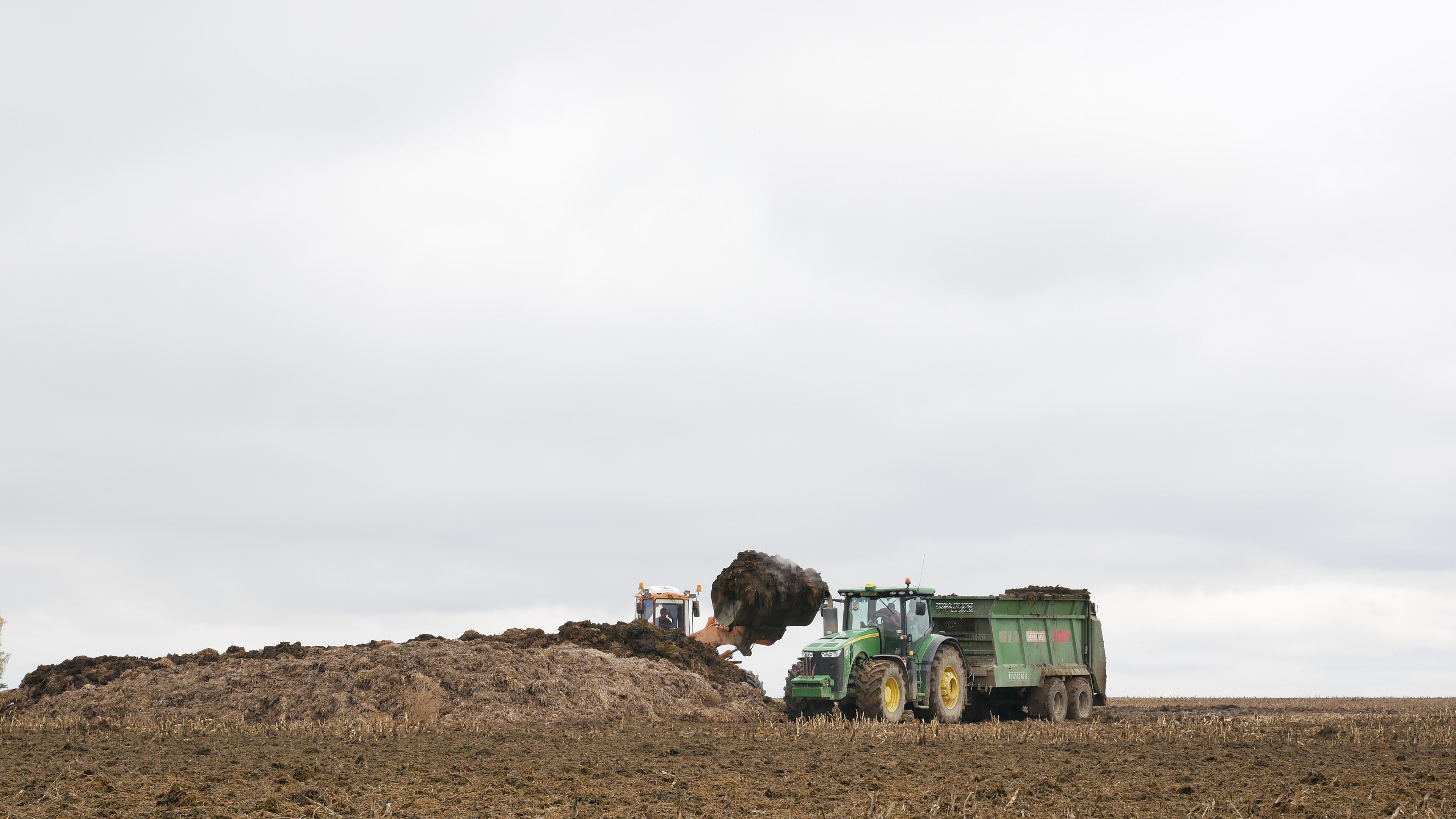 Pala cargadora llenando un remolque esparcidor de estiércol sólido desde un montón acopiado en una parcela, con el tractor verde enganchado y el campo labrado en primer plano