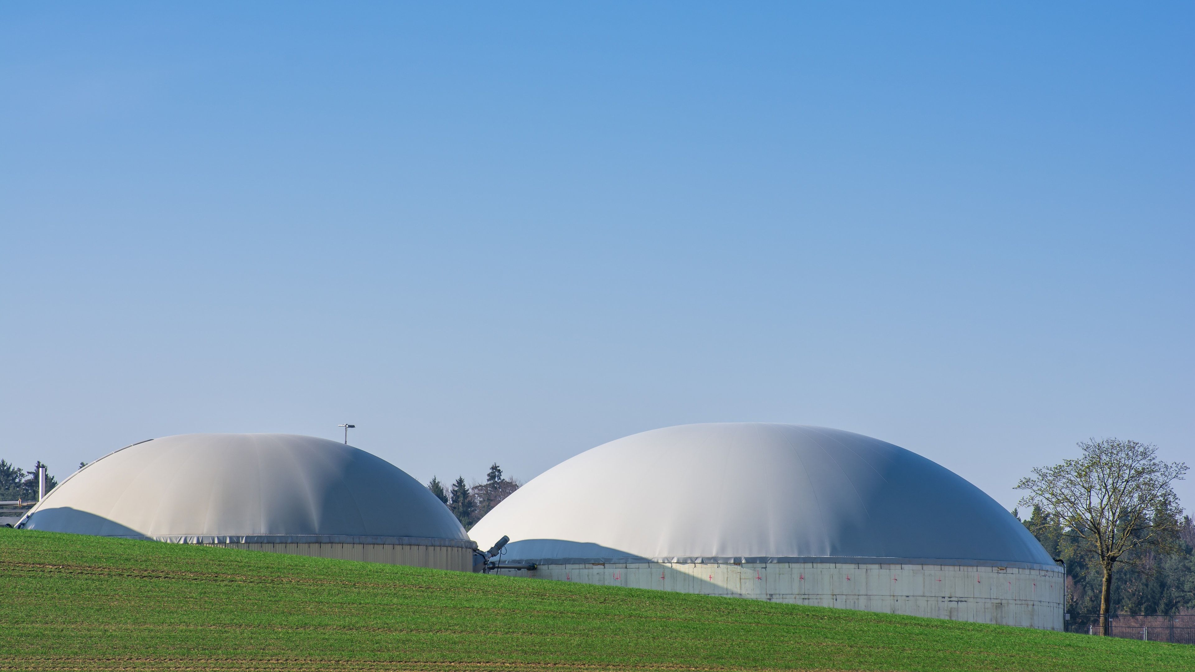 Dos digestores de biogás con cúpulas de membrana blancas sobre tanques de hormigón, con un agitador y una tubería entre ambos, junto a un campo sembrado