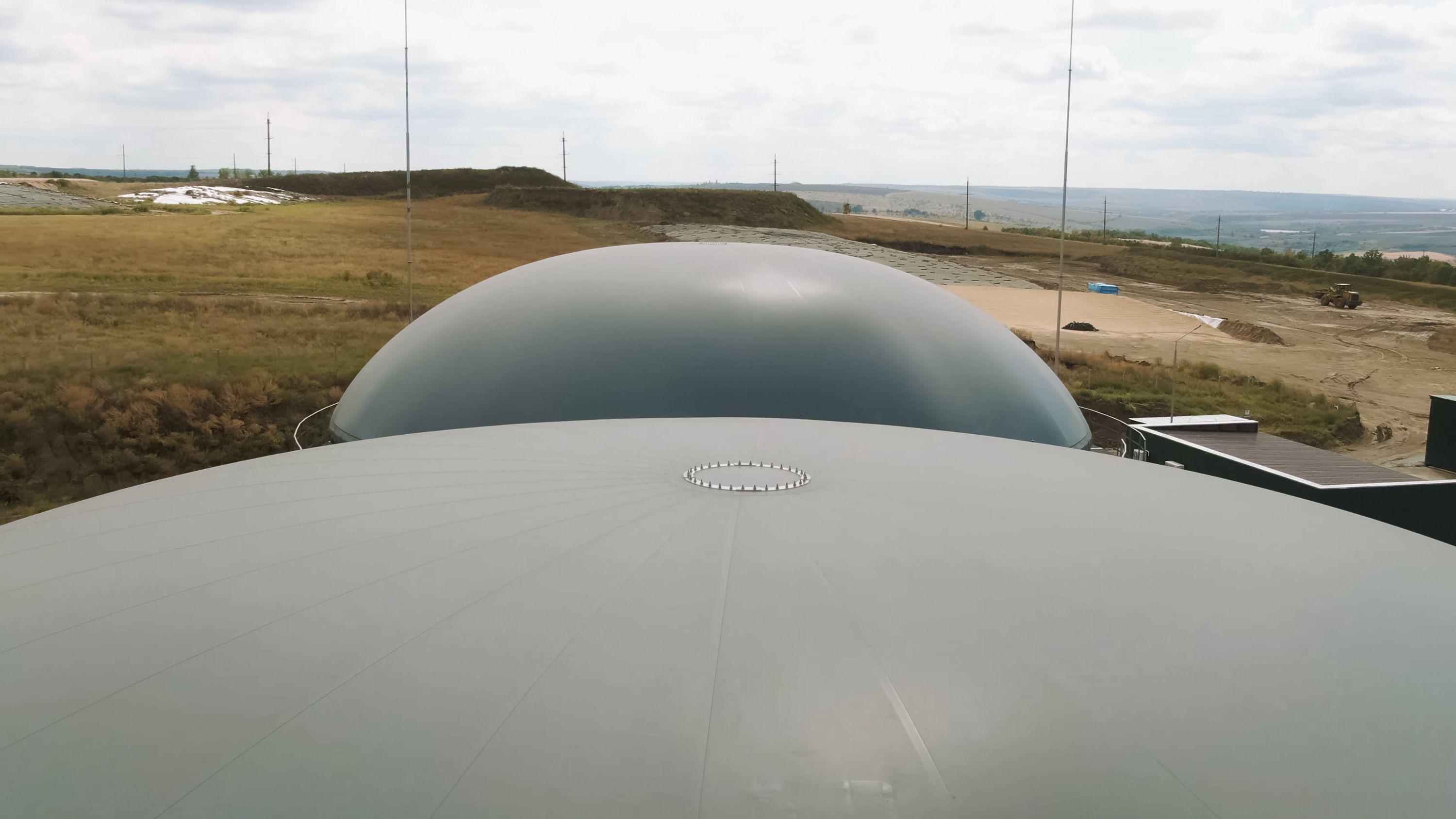 Cubiertas de membrana de dos digestores vistas desde lo alto; al fondo, la parcela todavía en obra, con terreno explanado y una pala cargadora trabajando