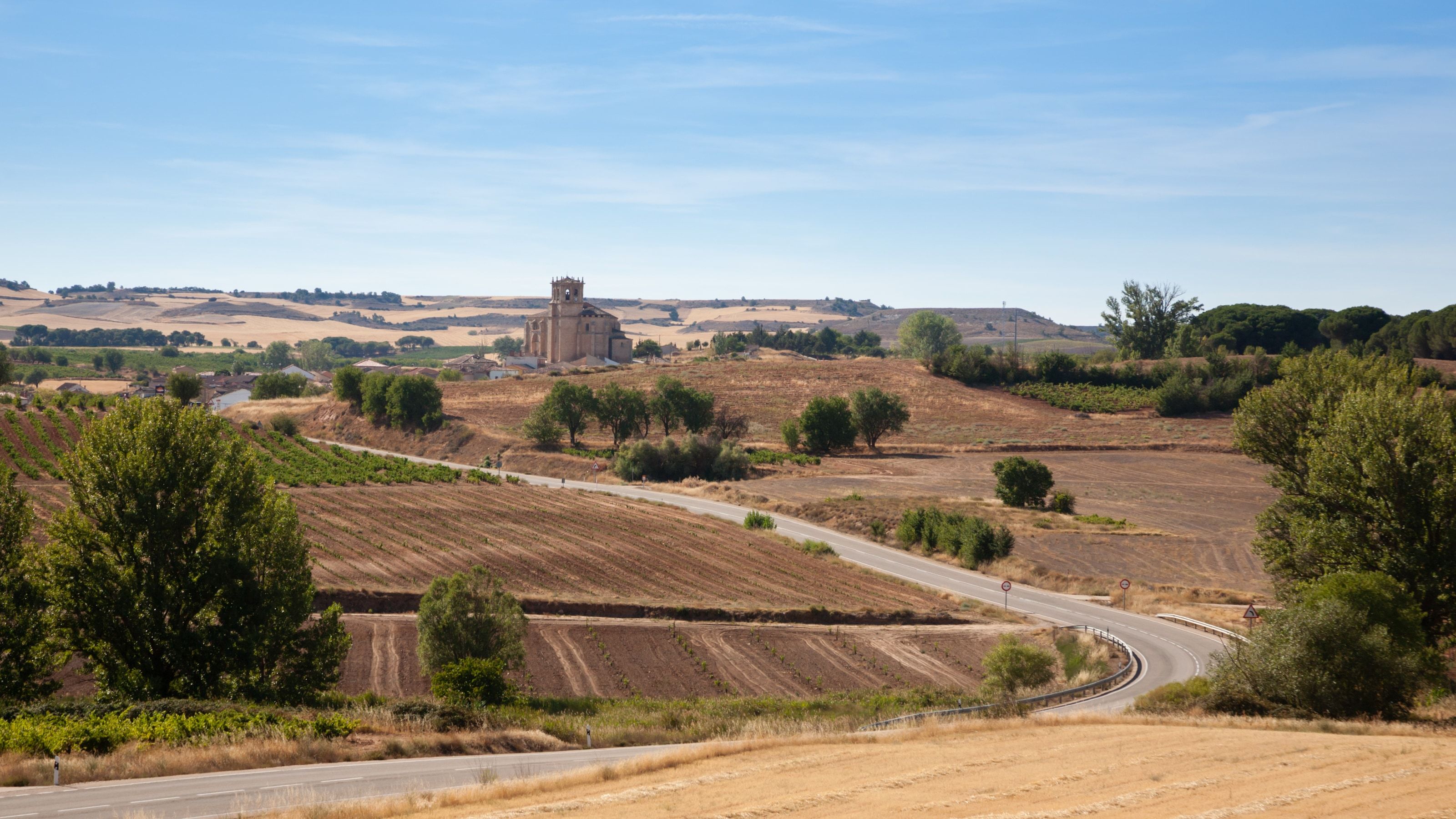 Paisaje agrícola de secano en Castilla y León con parcelas aradas, una carretera comarcal y un pueblo con iglesia al fondo
