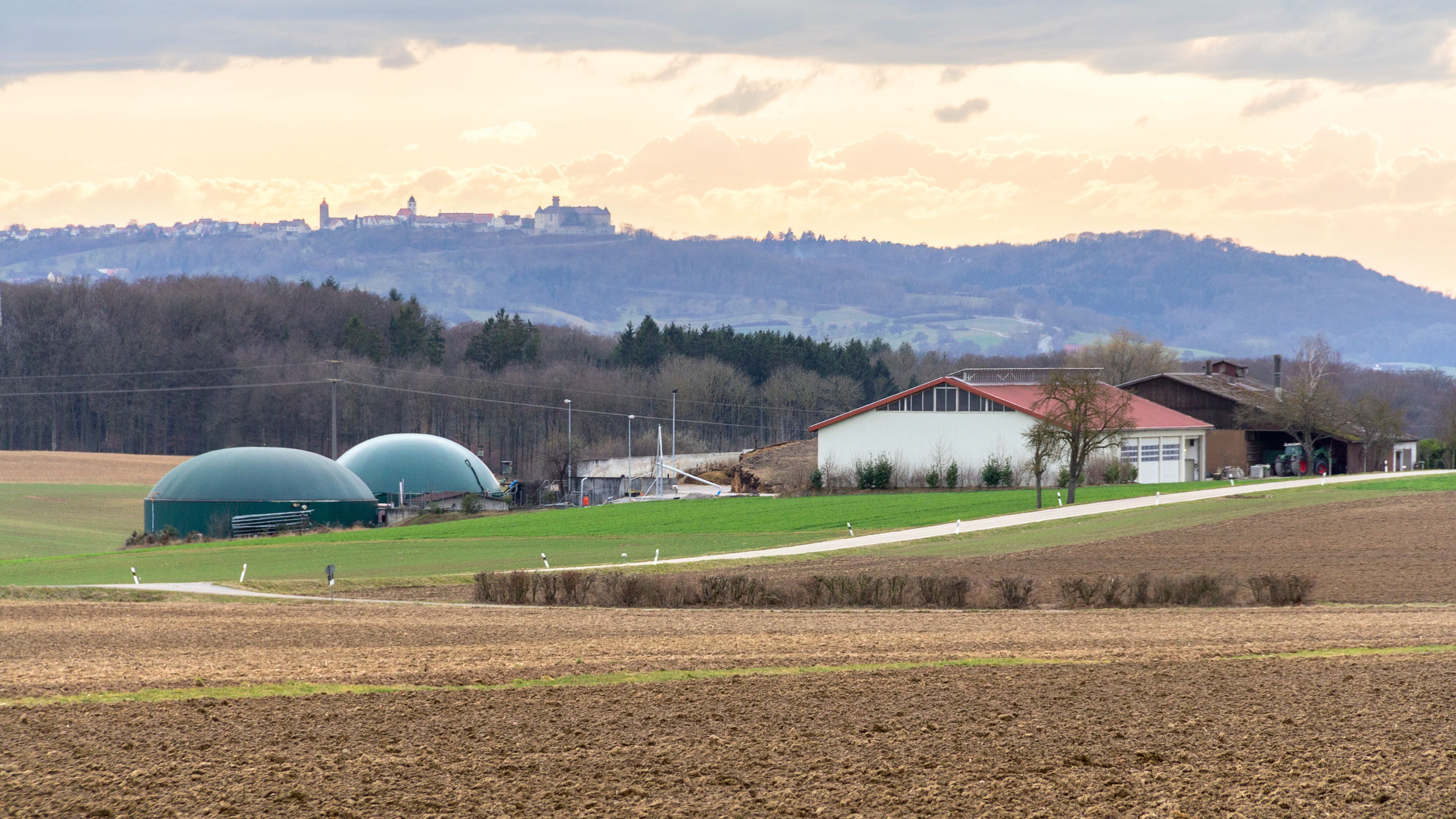 Planta de biogás de una explotación agrícola: dos digestores de cubierta verde junto a una nave de tejado rojo, con un campo arado en primer plano y un pueblo con castillo en la colina del fondo